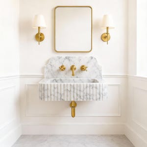 Puede incluir: Conjunto de ba&ntilde;o con un lavabo de m&aacute;rmol con grifos dorados, un espejo rectangular con marco dorado y dos apliques de pared con pantallas blancas. El lavabo tiene un dise&ntilde;o acanalado y est&aacute; montado en una pared blanca.