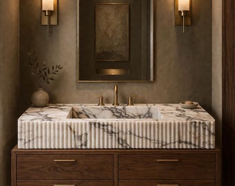 May include: A modern bathroom vanity with a marble sink and a dark wood cabinet. The sink has a ribbed design and gold fixtures. Two sconces flank a large mirror. A small vase with greenery sits on the counter.