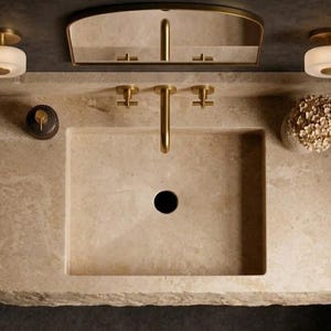 Puede incluir: Un lavabo rectangular de piedra beige con un grifo de lat&oacute;n y dos manijas a juego. El conjunto incluye un espejo con marco de lat&oacute;n, dos apliques redondos de vidrio esmerilado, un dispensador de jab&oacute;n oscuro y un arreglo floral blanco.
