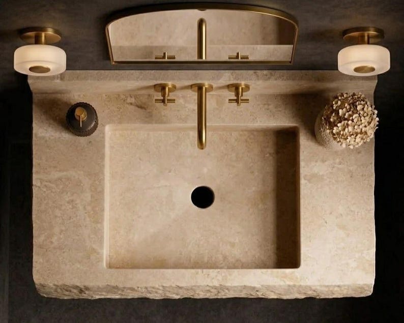 Puede incluir: Un lavabo rectangular de piedra beige con un grifo de lat&oacute;n y dos manijas a juego. El conjunto incluye un espejo con marco de lat&oacute;n, dos apliques redondos de vidrio esmerilado, un dispensador de jab&oacute;n oscuro y un arreglo floral blanco.