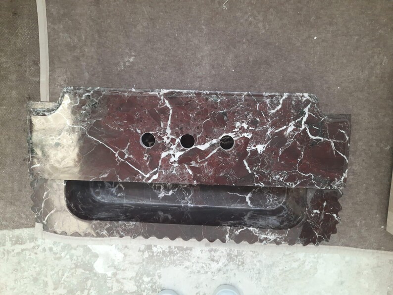 May include: A rectangular, dark red marble sink with white veining. The sink has three circular faucet holes and a built-in, rectangular basin. The edges are scalloped and the overall design is modern.