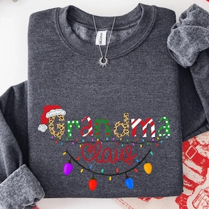 May include: A dark grey sweatshirt featuring the words "Grandma Claus" in a festive design. "Grandma" is in leopard print, "Claus" in red and green. Multicoloured Christmas lights are below the text, creating a holiday theme.
