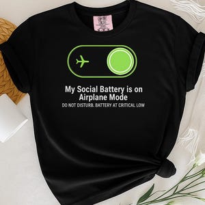 Puede incluir: Camiseta negra con el texto "My Social Battery is on Airplane Mode" y un icono de avi&oacute;n verde. La camiseta tambi&eacute;n dice "DO NOT DISTURB. BATTERY AT CRITICAL LOW". La camiseta se muestra sobre una superficie blanca con una taza blanca y flores.