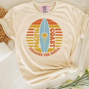 May include: Cream-colored t-shirt featuring a circular graphic with a surfboard, palm tree, and the words "Pacific Coast Chasing the Swell" in orange, yellow, and blue. The shirt is styled with a woven basket, a white mug, and flowers.