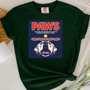 May include: Dark green t-shirt featuring a graphic with the word "PAWS" in red, a dog illustration, and the text "JUST WHEN YOU THOUGHT IT WAS SAFE TO GO BACK IN THE WATER... FETCH." A white mug and a woven mat are visible.