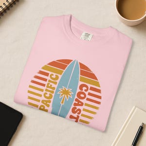 May include: A light pink t-shirt featuring a retro design with a surfboard and palm tree graphic. The design includes the words "Pacific Coast" in a circular arrangement, with orange, yellow, and blue colors.