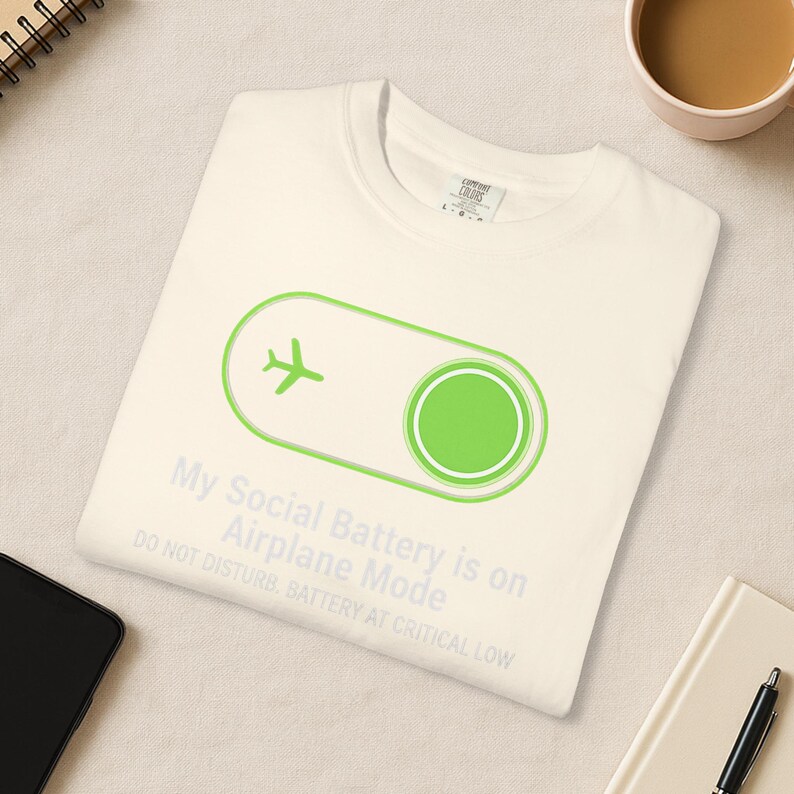 Puede incluir: Camiseta color crema con un icono de avi&oacute;n verde y el texto "My Social Battery is on Airplane Mode. Do Not Disturb. Battery at Critical Low." La camiseta est&aacute; doblada sobre una superficie clara.