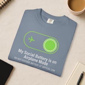 Puede incluir: Una camiseta azul pizarra con un gr&aacute;fico verde de modo avi&oacute;n y el texto "My Social Battery is on Airplane Mode. Do Not Disturb. Battery at Critical Low." La camiseta est&aacute; doblada.