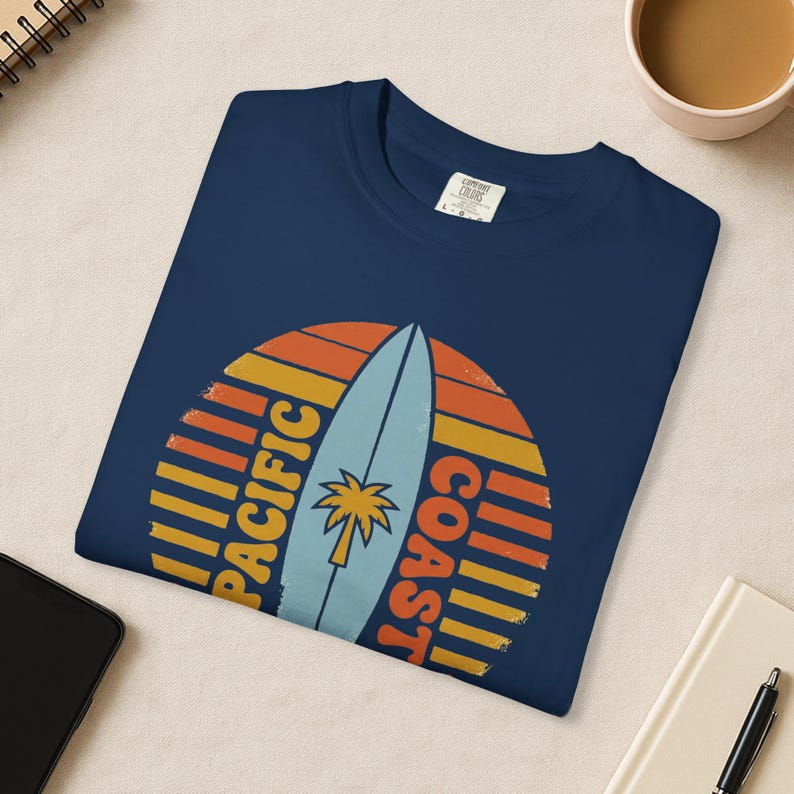 May include: Navy blue t-shirt featuring a retro design with a surfboard and palm tree. The design includes the words "PACIFIC COAST" in orange and yellow, set against a sunset-style background.