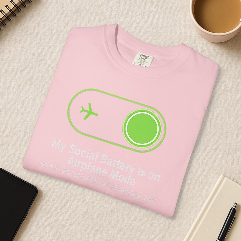 Puede incluir: Camiseta rosa claro con el texto "My Social Battery is on Airplane Mode" y un icono de avi&oacute;n verde. La camiseta tambi&eacute;n tiene un dise&ntilde;o de interruptor basculante verde. El texto "DO NOT DISTURB BATTERY AT CRITICAL LOW" tambi&eacute;n est&aacute; impreso en la camiseta.