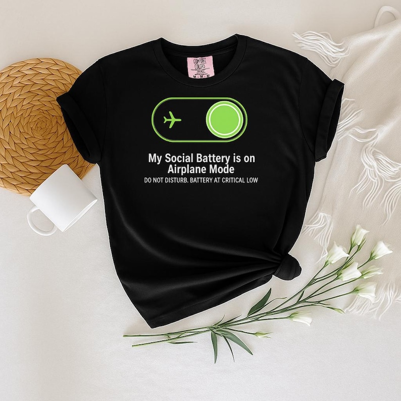 Puede incluir: Camiseta negra con el texto "My Social Battery is on Airplane Mode" y un icono de avi&oacute;n verde. La camiseta tambi&eacute;n dice "DO NOT DISTURB. BATTERY AT CRITICAL LOW". La camiseta se muestra sobre una superficie blanca con una taza blanca y flores.