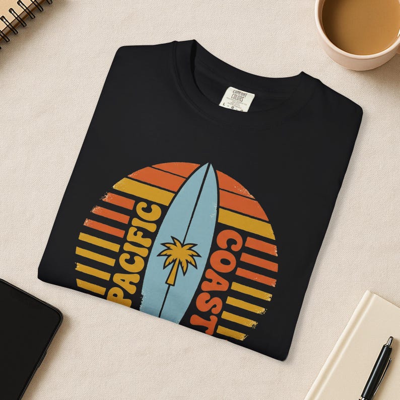 May include: Black t-shirt featuring a retro design with a surfboard and palm tree. The design includes the words "PACIFIC COAST" in a vintage font, set against a sunset-style background. A casual, beach-themed apparel item.