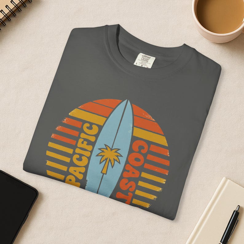 May include: Dark gray t-shirt with a retro design featuring a light blue surfboard and a palm tree, set against a sunset-style background. The words "PACIFIC COAST" are printed on the shirt.