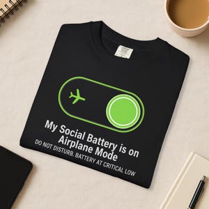 Puede incluir: Camiseta negra con un gr&aacute;fico verde de modo avi&oacute;n y el texto "My Social Battery is on Airplane Mode. Do Not Disturb. Battery at Critical Low." La camiseta est&aacute; doblada sobre una superficie beige.