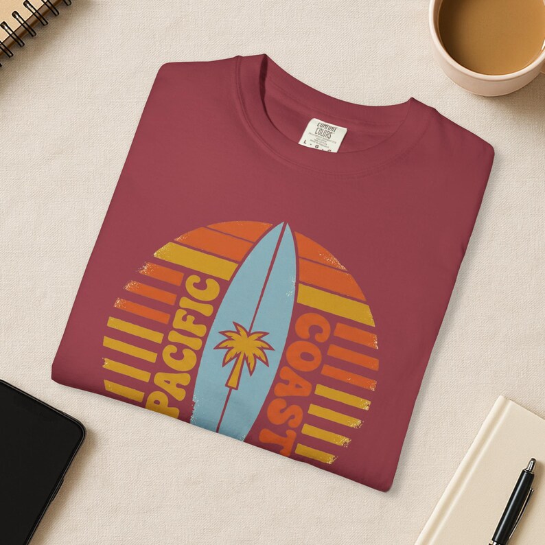 May include: A maroon t-shirt featuring a retro design with a surfboard and palm tree. The design includes the words "PACIFIC COAST" in a vertical layout, set against a sunset-style background with orange and yellow stripes.