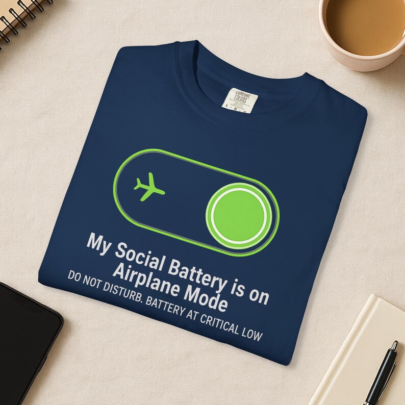 Puede incluir: Camiseta azul marino con un gr&aacute;fico verde y blanco de un avi&oacute;n y un bot&oacute;n de encendido. El texto dice: "Mi bater&iacute;a social est&aacute; en modo avi&oacute;n. No molestar. Bater&iacute;a baja cr&iacute;tica."