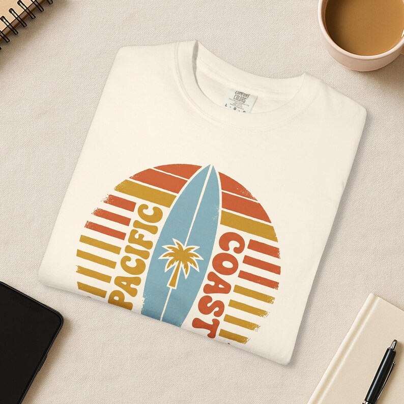 May include: Cream-colored t-shirt featuring a retro design with a surfboard and palm tree. The graphic includes the words "Pacific Coast" in orange and yellow, set against a sunset-style background. The shirt is folded neatly.