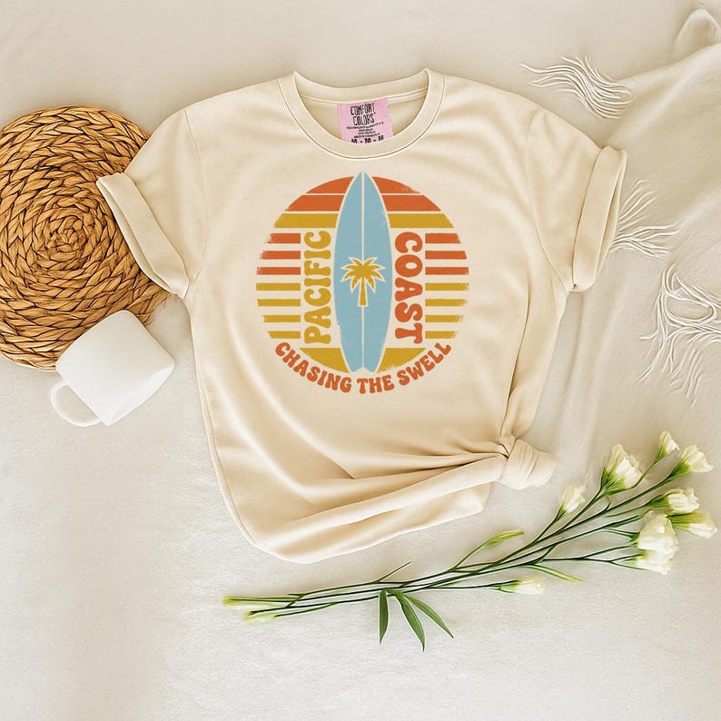 May include: Cream-colored t-shirt featuring a circular graphic with a surfboard, palm tree, and the words "Pacific Coast Chasing the Swell" in orange, yellow, and blue. The shirt is styled with a woven basket, a white mug, and flowers.
