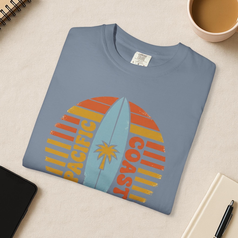 May include: A blue t-shirt with a graphic featuring a surfboard and a palm tree, set against a sunset design. The text "PACIFIC COAST" is printed in yellow and orange. The shirt is folded neatly.