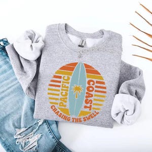 May include: A gray sweatshirt with a retro design featuring a surfboard, palm tree, and the words "Pacific Coast" and "Chasing the Swell" in orange, yellow, and blue. The sweatshirt is paired with light blue jeans.
