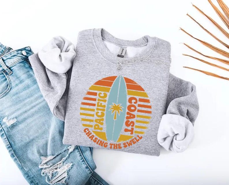 May include: A gray sweatshirt with a retro design featuring a surfboard, palm tree, and the words "Pacific Coast" and "Chasing the Swell" in orange, yellow, and blue. The sweatshirt is paired with light blue jeans.