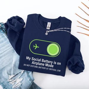 Puede incluir: Sudadera azul marino con el texto "My Social Battery is on Airplane Mode" y un avi&oacute;n verde. El dise&ntilde;o incluye un bot&oacute;n de encendido verde y la frase "DO NOT DISTURB. BATTERY AT CRITICAL LOW."