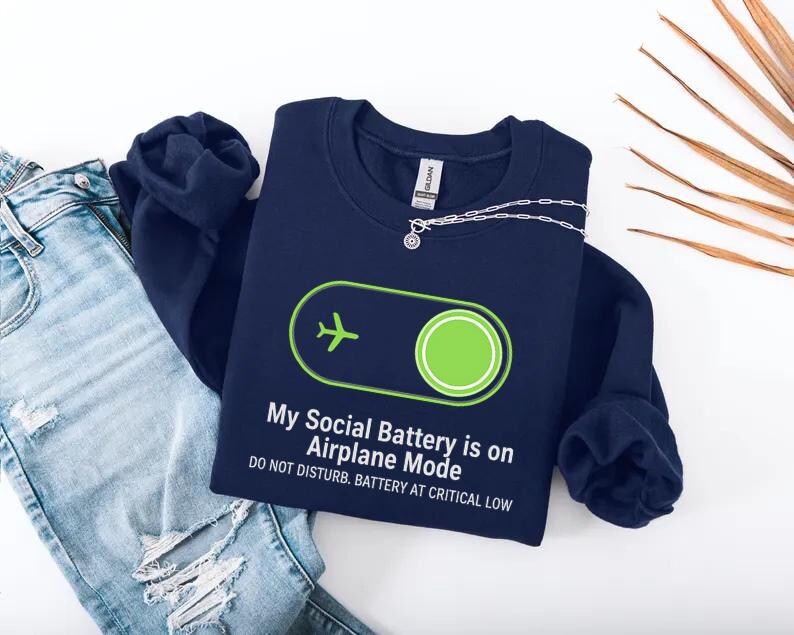 Puede incluir: Sudadera azul marino con el texto "My Social Battery is on Airplane Mode" y un avi&oacute;n verde. El dise&ntilde;o incluye un bot&oacute;n de encendido verde y la frase "DO NOT DISTURB. BATTERY AT CRITICAL LOW."