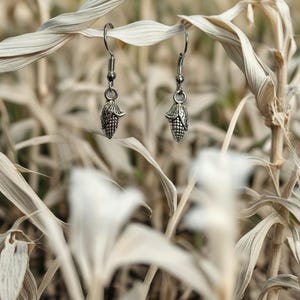 May include: Silver-toned corn-shaped earrings hanging from a dried plant stalk. The earrings have a detailed texture, resembling miniature corn cobs. The background is a blurred field of dry plants, creating a rustic, natural aesthetic.