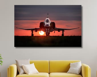 May include: A framed canvas print of a jet airplane on a runway at sunset. The sun is centered behind the plane, creating a silhouette effect. The sky is a gradient of orange and purple.