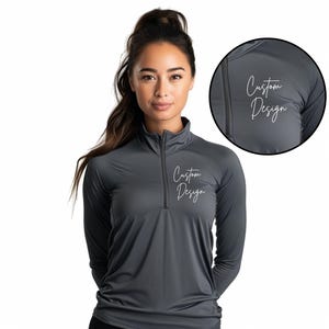 May include: A charcoal gray long-sleeve athletic shirt with a quarter-zip collar. The shirt features the text "Custom Design" in a white script font on the chest. The image shows a close-up of the shirt's fabric and design.