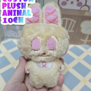 May include: A plush bunny toy in a light tan color with pink accents. The bunny has embroidered pink eyes and ears, and a small bib with a cross design. The text "CUSTOM PLUSH ANIMAL 10CM" is visible.
