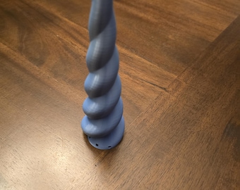 Peut inclure: Une corne de licorne bleu clair en spirale. La corne est de couleur unie et a un aspect texturé. La base comporte trois petits trous. Elle est posée sur une surface en bois.