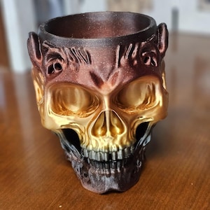 May include: A decorative skull-shaped container with a metallic gold and bronze finish. The skull features detailed facial features and a wide opening at the top. The container is likely used for holding small items or as a unique decorative piece.