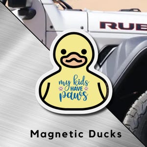 Op de afbeelding: Een gele eendvormige magneet met een zwarte omtrek en een cartoongezicht. De tekst "my kids have paws" is in blauwe letters geschreven. De magneet wordt weergegeven tegen een zilveren achtergrond met een wit voertuig op de achtergrond.