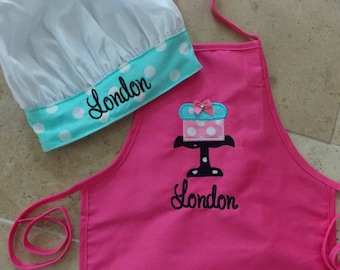 Peut inclure: Un ensemble tablier et toque de chef rose pour enfant. Le tablier présente un motif de gâteau et le nom "London" brodé en noir. La toque est blanche avec une bande à pois turquoise et le nom "London" brodé en noir.