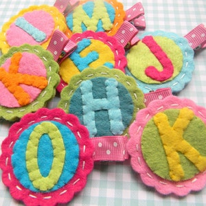 May include: Nine felt circles with different letters of the alphabet, each with a pink alligator clip attached. The circles are in various colors, including pink, green, yellow, and blue.