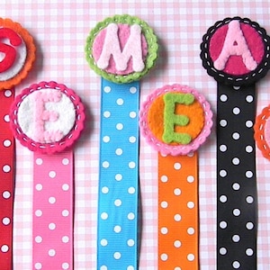 May include: A collection of colorful handmade felt bookmarks. Each bookmark features a ribbon with white polka dots and a felt circle with a letter. The letters spell out a word, with each letter in a different color.