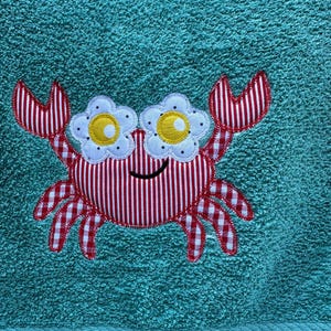 May include: A teal towel with a crab applique. The crab is red and white gingham with red and white striped claws. The crab's eyes are white flowers with yellow centers. The crab has a smiling black mouth.