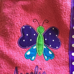 May include: Pink hooded towel with a butterfly applique. The butterfly has purple wings with white polka dots, blue accents, and a green body. The name "Amelia" is embroidered in purple script.