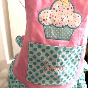 May include: A pink apron featuring a large cupcake appliqué with a yellow crocheted flower on top. The cupcake has a white top with colorful polka dots and a light blue polka dot base. A matching light blue polka dot pocket displays the name Gracie.