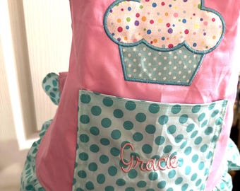 May include: A pink apron featuring a large cupcake appliqué with a yellow crocheted flower on top. The cupcake has a white top with colorful polka dots and a light blue polka dot base. A matching light blue polka dot pocket displays the name Gracie.