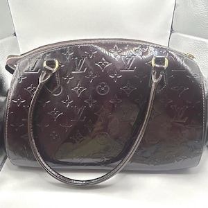 May include: A glossy, dark brown handbag with embossed patterns and gold-toned hardware. The bag features a zip closure and two curved handles. The surface reflects light, highlighting the embossed design.