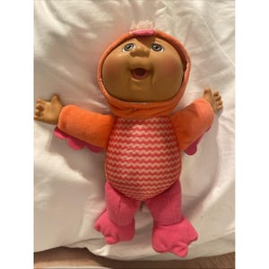 May include: A Cabbage Patch Kids doll wearing an orange hooded outfit with pink accents. The doll has a round face, rosy cheeks, and open mouth. The outfit has a pink and white zigzag pattern on the torso and pink feet.