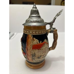 May include: A ceramic beer stein with a detailed scene of a house with a red roof and mountains. The stein has a metal lid and handle. The word "Timmer" is printed on the base of the stein.