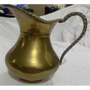 Könnte beinhalten: Eine Vintage-Messingkanne mit gebogenem Griff und dekorativem Rand. Die Kanne hat einen bauchigen Körper und eine polierte Oberfläche, die das Licht reflektiert. Der Griff ist gedreht und verleiht dem Stück ein einzigartiges Detail.
