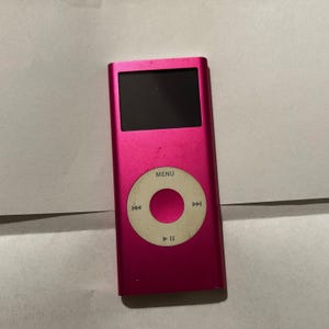 May include: A hot pink digital music player with a rectangular black screen and a circular white click wheel. The click wheel features the word MENU at the top, along with symbols for play, pause, and track skipping. The device has a metallic finish.