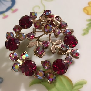 Op de afbeelding: Een vintage goudkleurige broche met een bloemmotief. De broche heeft een cirkelvormige opstelling van grote, ronde, dieprode edelstenen en kleinere iriserende stenen, met goudkleurige bladeren en stelen.