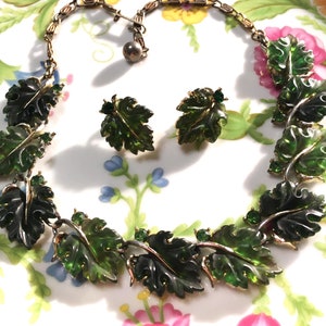 May include: A gold-tone metal necklace and matching clip-on earrings set featuring translucent dark green leaf-shaped components. The necklace consists of linked leaf segments, while the earrings are single leaf designs with small green accents.