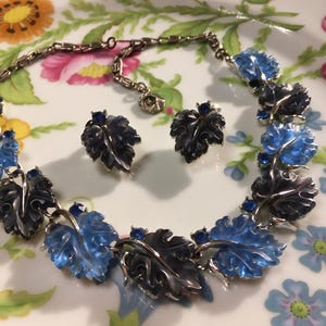 May include: A vintage necklace and matching earrings set. The necklace and earrings feature leaf-shaped pendants in shades of blue and black, accented with small blue gemstones. The jewelry is set against a floral background.