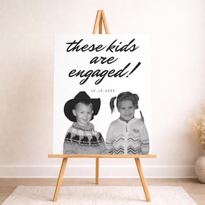 Op de afbeelding: Een zwart-wit verlovingsaankondigingsbord op een houten ezel. Het bord leest "these kids are engaged!" met de datum "10.19.2025" eronder. Het bord bevat twee vintage foto's van kinderen.